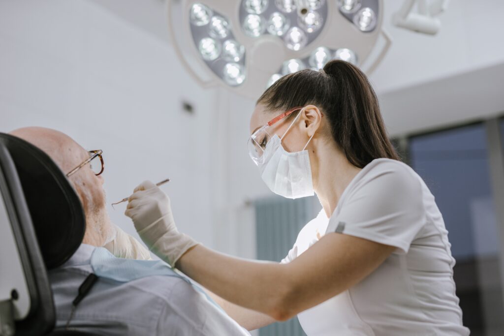 Female dentist in a mask and gloves examining an older patient's teeth in a bright, modern dental office