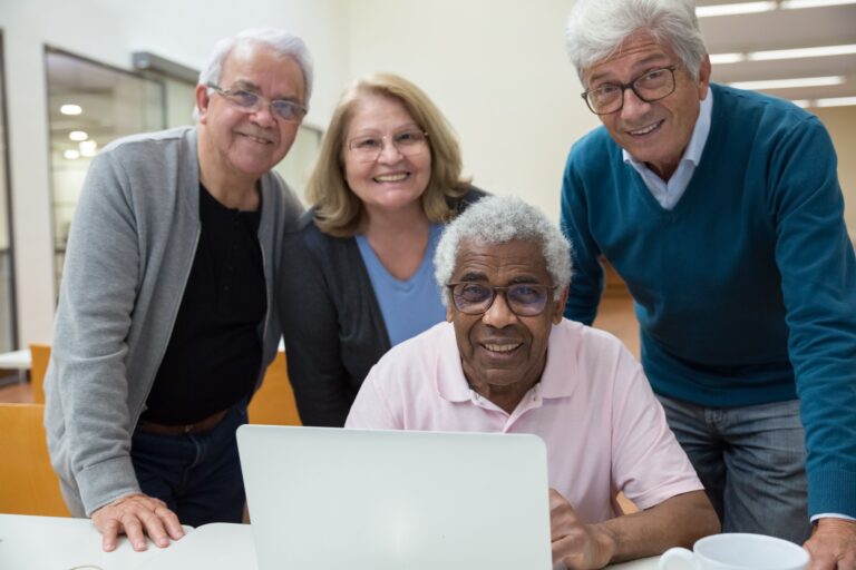 Four smiling senior adults gathered around a laptop in a classroom, learning Medicare basics together