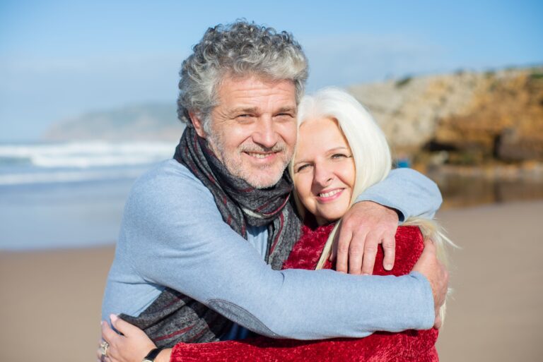 Smiling older couple hugging on a sunny beach, at ease after sorting out their Medicare Supplement open enrollment questions.
